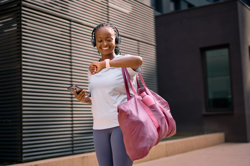 African American woman smiling while checking her fitness tracker, holding a smartphone and a gym bag, representing a healthy and active urban lifestyle