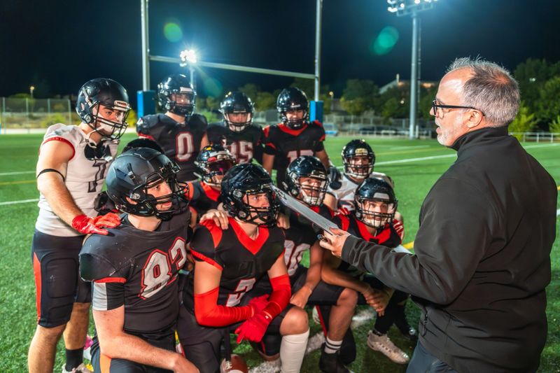American football coach instructing young adult players in a focused night practice huddle on a brightly lit stadium field, planning strategy and team execution