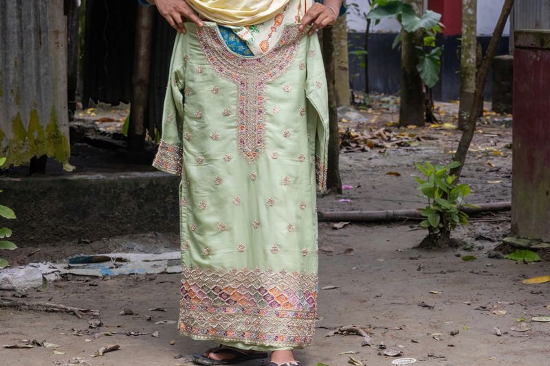 A Bangladeshi woman's hand presenting a highly decorated Kameez with colorful beads and embroidery. Traditional South Asian fashion, ethnic dress, and textile craft in a village setting.
