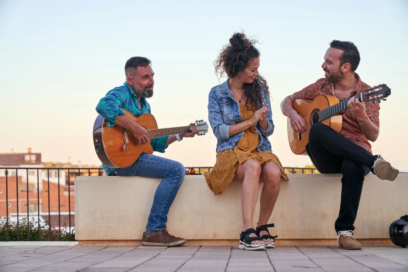 Three friends, two men playing acoustic guitars and a woman clapping, enjoying a music session on a rooftop
