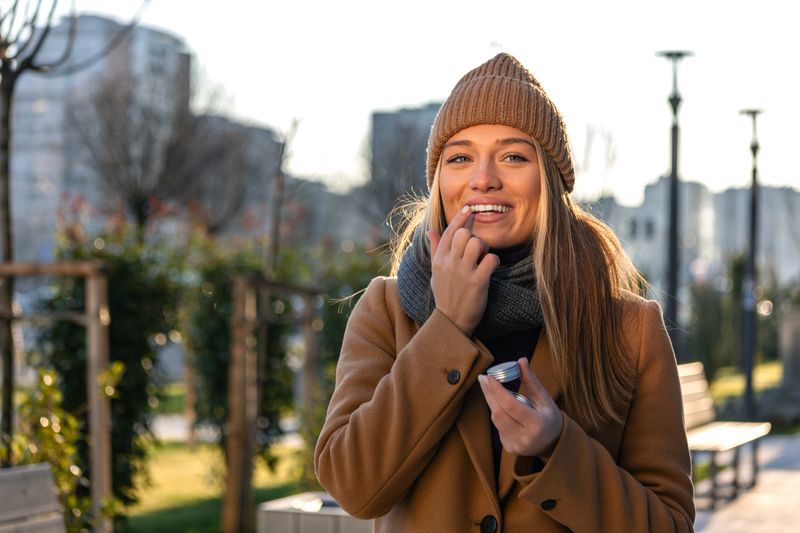 Happy young woman in a brown coat and knitted beanie applying lip balm outdoors on a Cold Weather. Concept of skincare, beauty, hydration, and winter self-care routine.