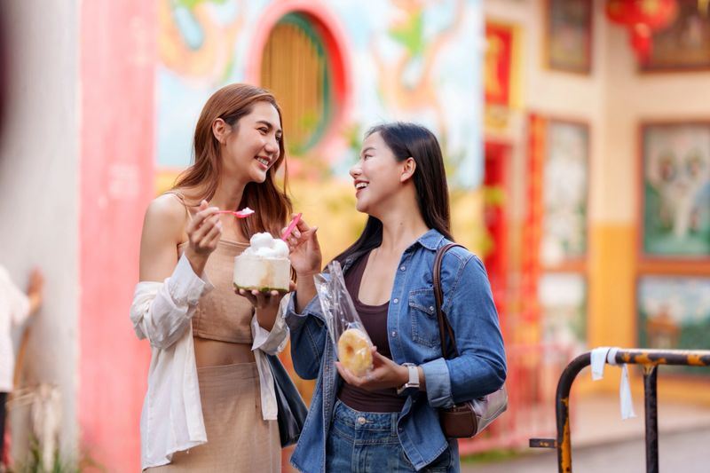 Two cheerful young adult asian women friends eating street food while smiling and exploring a vibrant market during vacation