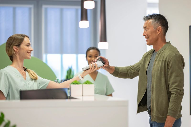 Mature Asian male smiling while paying at a physical therapy clinic featuring modern decor and friendly staff. He wears a casual green jacket and uses a card reader, interacting positively with the receptionist.