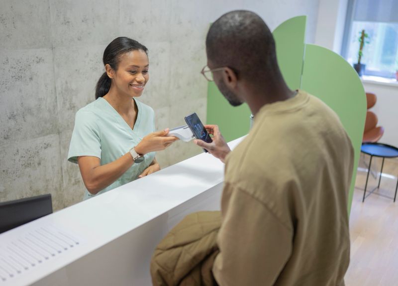 Mid adult Black male paying at a physical therapy clinic reception featuring a smiling receptionist in scrubs. The setting includes a modern reception area with a card payment being processed, indicating a professional healthcare environment.
