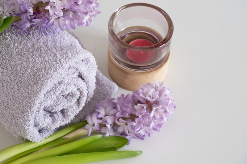 A rolled lavender-colored towel with delicate purple hyacinth flowers and a wooden candle holder with a candle, arranged on a clean white surface. The soft textures and pastel tones create a relaxing and inviting atmosphere, ideal for spa, wellness, and self-care concepts. Captured from a slightly elevated angle, emphasizing the details of the towel's texture and flower petals. Ideal for spa and wellness advertising, beauty product promotions, relaxation and self-care themes, social media posts about relaxation, aromatherapy, or home decor
