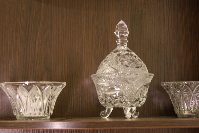 Elegant items made of glass or crystal are displayed on a wooden cabinet shelf, including a figured sugar bowl with a lid and two vases.