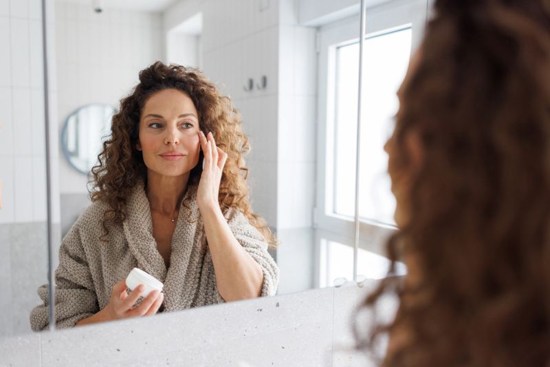 A mid adult woman with curly hair wearing a cozy bathrobe applies face cream to her face in a bright, modern bathroom with natural daylight streaming through the window.