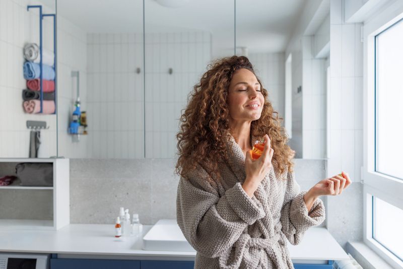 A mid adult woman with curly hair wearing a cozy robe applies fragrance in a bright modern bathroom with white tiles and natural daylight.