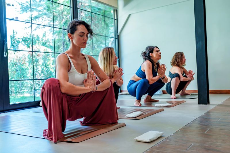A group of women practicing a seated twist yoga pose in a warm, inviting studio with a wooden floor, brown yoga mats, and a rustic wooden table adorned with plants. The setting includes natural light from a window, colorful leggings, and scattered props like blankets and water bottles, creating a vibrant yet relaxed atmosphere. This high-resolution stock photo is perfect for fitness blogs, yoga class promotions, wellness retreats, or content on healthy lifestyles and community exercise.