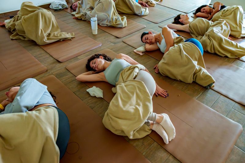 A group of people relaxing under blankets during a guided meditation session in a serene yoga studio. The scene features participants with eye masks, cozy mats, and a calming environment with natural light, perfect for wellness, stress relief, and mindfulness content. High-resolution stock photo ideal for health blogs, spa advertising, or mental health resources