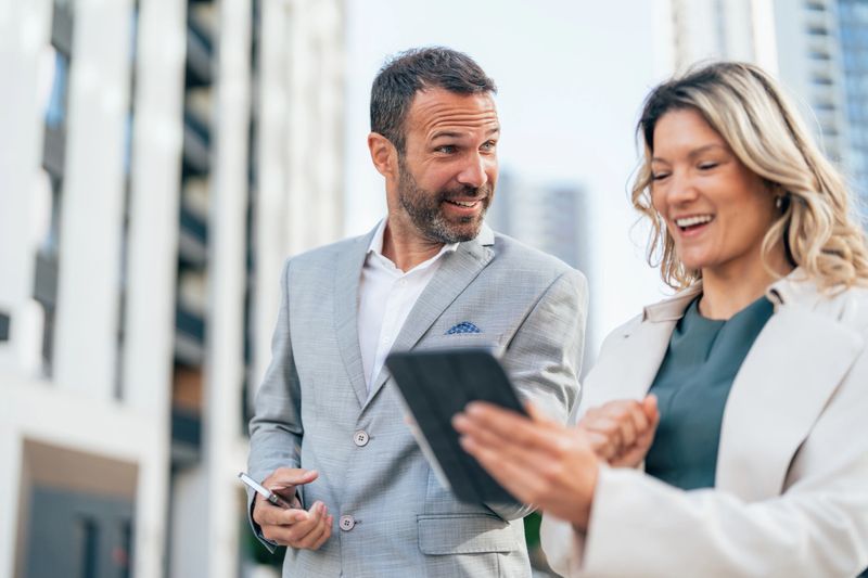 Shot of two confident colleagues walking and talking next to an office building. Businessman and businesswoman in meeting using digital tablet and discussing business strategy. Creative business persons discussing new project and sharing ideas while walking in front of their office building.
