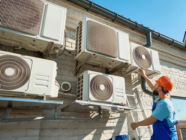 Technician on ladder servicing outdoor AC compressor units.