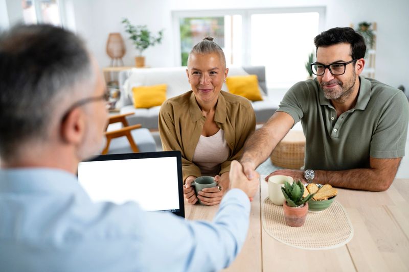 A financial advisor is shaking hands with a young man while a woman sits beside him holding a cup. They are in a bright, modern living room with cozy furniture and plants, suggesting a professional yet comfortable meeting environment. This image is suitable for themes related to financial planning, consulting, and family finance.
