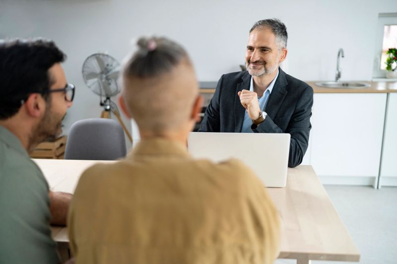 A professional financial advisor is engaged in a friendly meeting with a young family in a bright, modern kitchen. The advisor is smiling and appears to be explaining financial options or planning strategies, creating a positive and collaborative atmosphere. This image is suitable for financial services, consulting, and family financial planning themes.