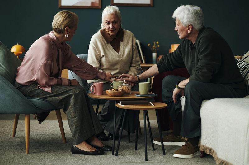 Three senior Caucasian people, women and man, sitting together, sharing snacks and drinks, engaging in conversation around small coffee tables in living room, relaxed body language