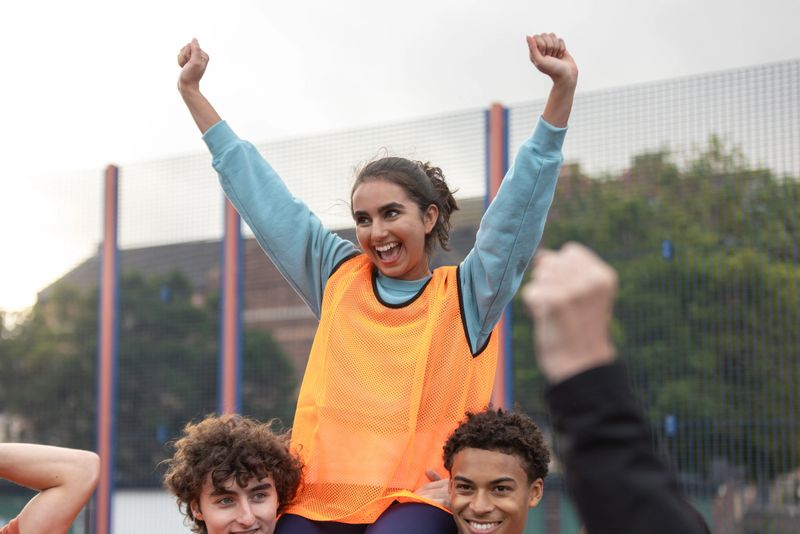 A medium view of a small group of teenagers celebrating in a public basketball court in Newcastle, England. Casual sportswear and bibs in an urban court setting emphasise youth lifestyle, fitness, and recreation themes. A young girl is being carried on her friend's shoulders as they celebrate winning a basketball game.Videos are available similar to this scenario.