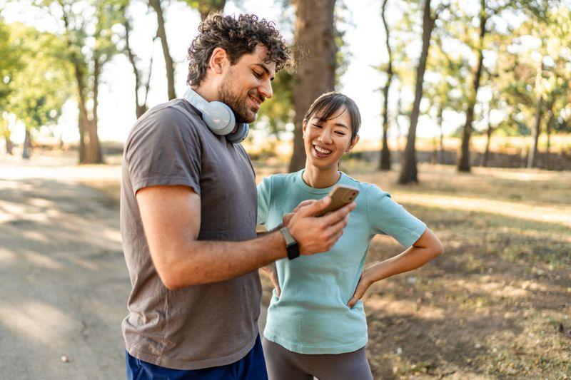Couple in athletic wear laughing and looking at smart phone after exercising outdoors, representing healthy lifestyle, digital connectivity, and shared fitness goals