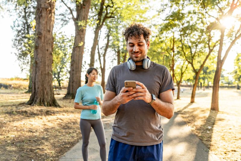 Young man in headphones checks his smartphone while chatting with a smiling partner holding a water bottle in a sunny park after exercise, relaxed and active outdoors