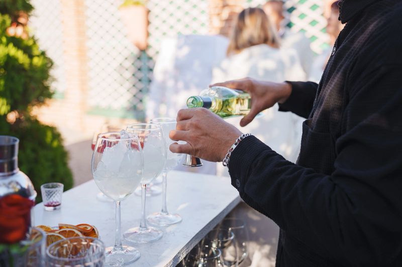A bartender carefully pours a beverage into a large glass at a lively outdoor event. Guests enjoy the atmosphere and socialize in the background, creating a festive mood.