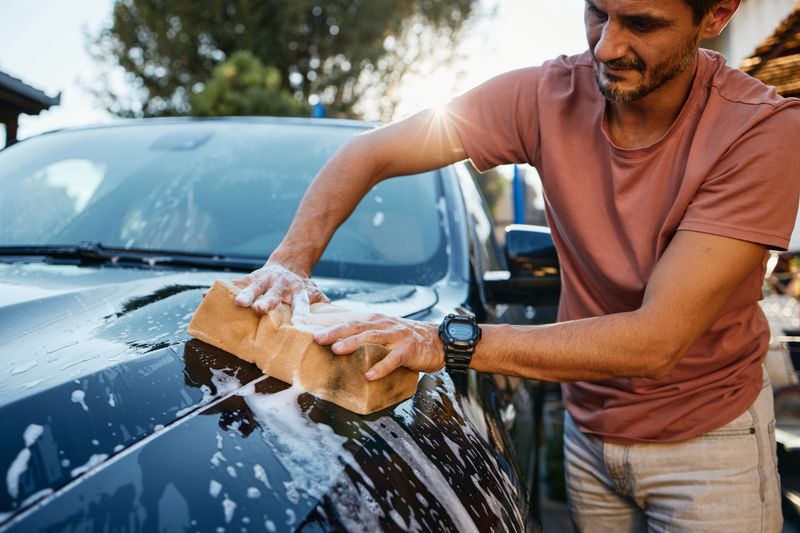 Young adult man carefully washing a black car with a sponge outdoors. Casual attire, focused expression, sunlight highlighting water and soap suds on the vehicle’s shiny surface. Summer or spring setting with greenery in the background.