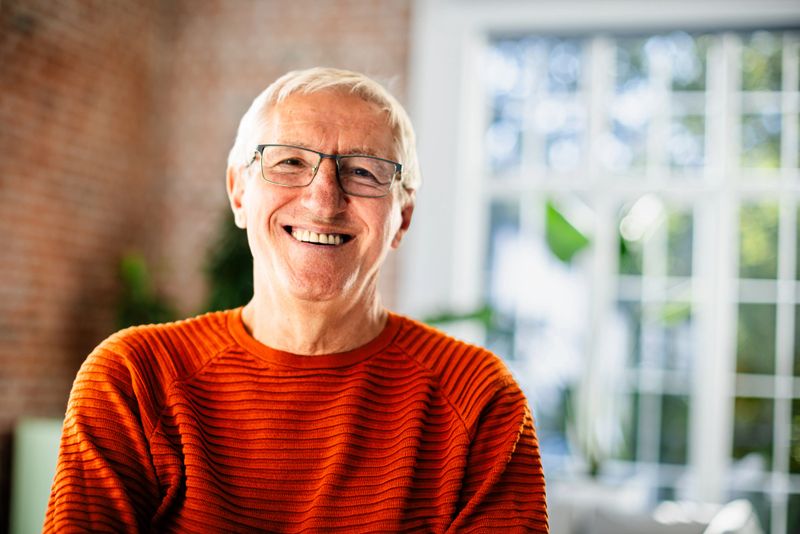 A cheerful senior man sits comfortably in his living room, dressed casually and wearing eyeglasses, enjoying his surroundings while radiating positivity and confidence.