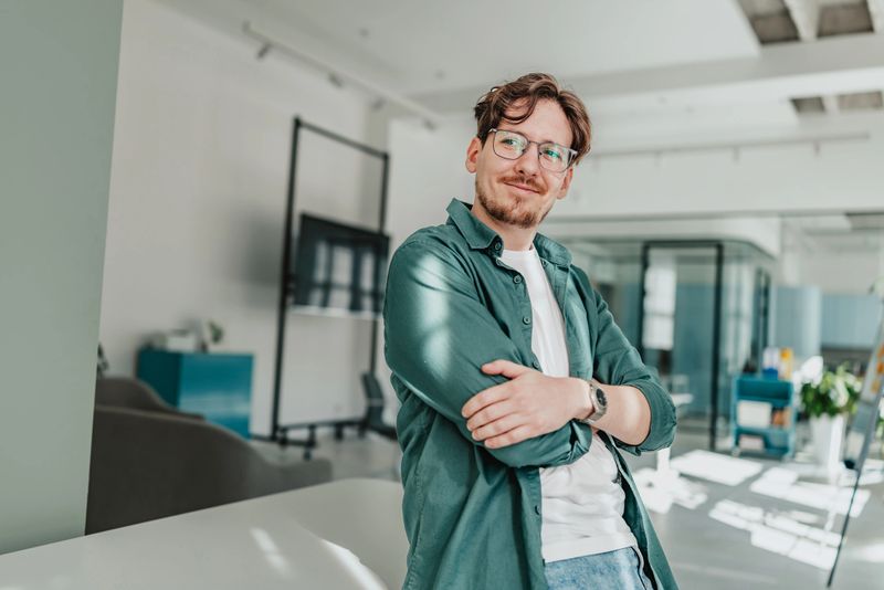 A young adult is smiling while standing confidently in a contemporary office. The workspace has a bright and inviting atmosphere, highlighting collaboration and enjoyment among colleagues.