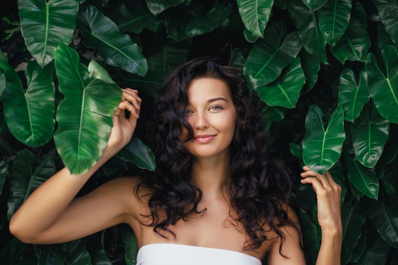 Beautiful woman with curly dark hair and glowing skin smiling in front of tropical greenery. Perfect for eco shampoo, natural beauty, skincare, and wellness product advertising.