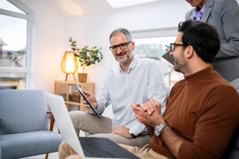 A woman stands behind two male colleagues seated on a couch in a bright office. They are slightly turned towards her, one holding a laptop on his lap and the other a tablet in his hands, as they engage in discussion and share ideas.