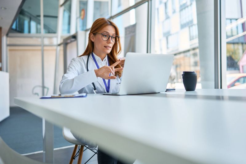 Female doctor in a white coat and glasses consulting a patient remotely, discussing medical advice and healthcare information during a virtual appointment using a laptop