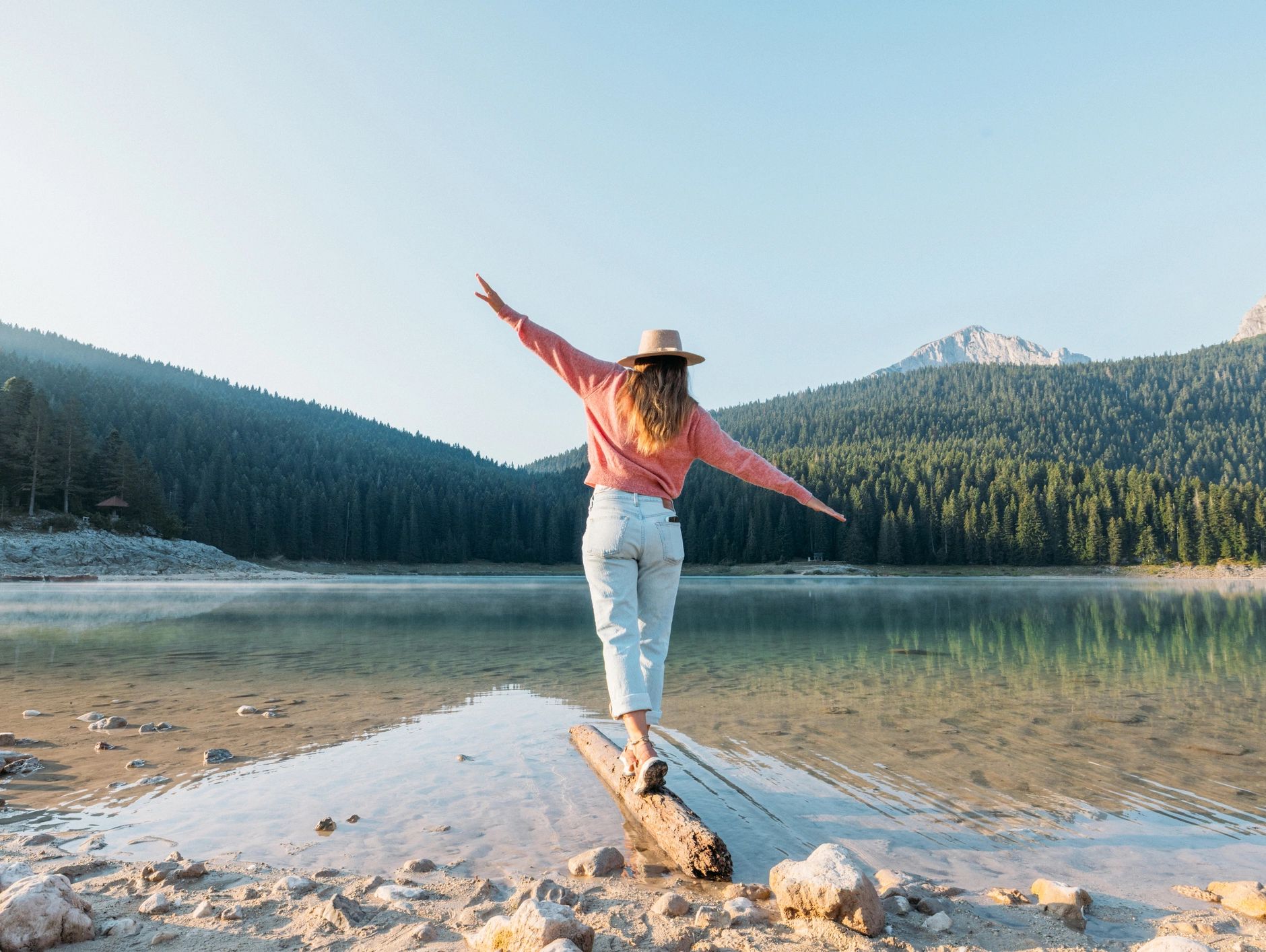 Woman balancing on a log by a serene lake surrounded by mountains.