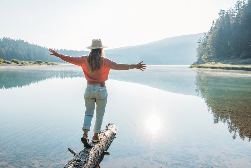 Young woman in hat and sweater balancing on a fallen log over a clear lake at sunrise. Outdoor adventure with serene water and forest background. Nature and mindfulness concept.