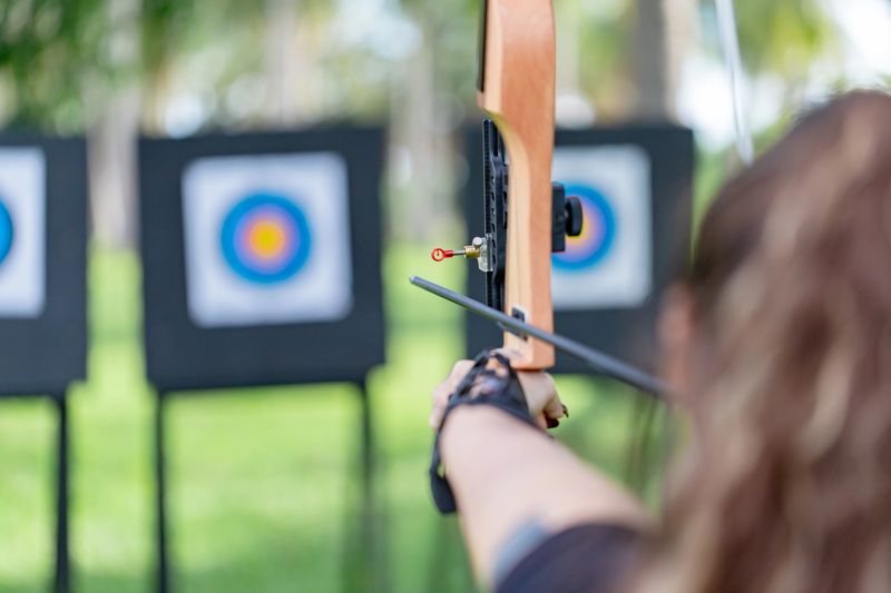 Female archer in black aiming her arrow intently at the bullseye on the outdoor range. A powerful image conveying goal achievement, determination, and sports training.