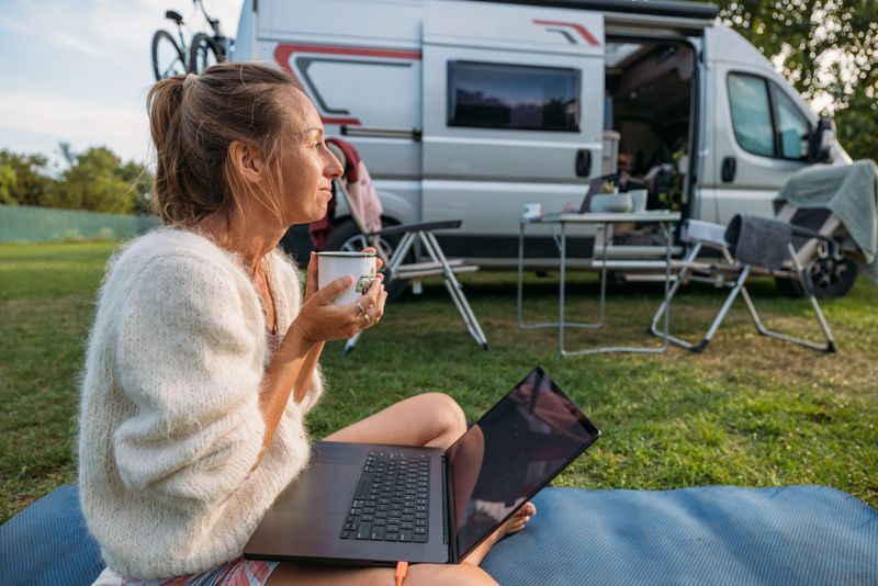 Adult woman sitting cross-legged on mat using laptop and holding mug outside camper van. Casual outdoor workspace with camping chairs and bike on van. Remote work lifestyle and nature.