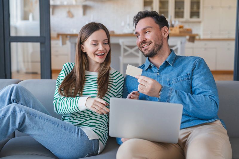 Credit card, laptop and couple on sofa in home with payment for hotel booking with holiday planning. Happy, computer and young man and woman with online banking app for vacation expenses at apartment