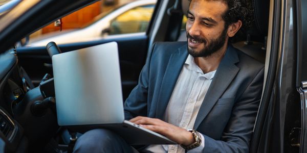Man in a suit working on a laptop inside a car, smiling.