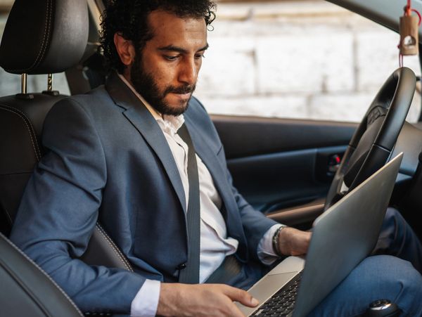 Man in suit using laptop inside a car, focused and serious.
