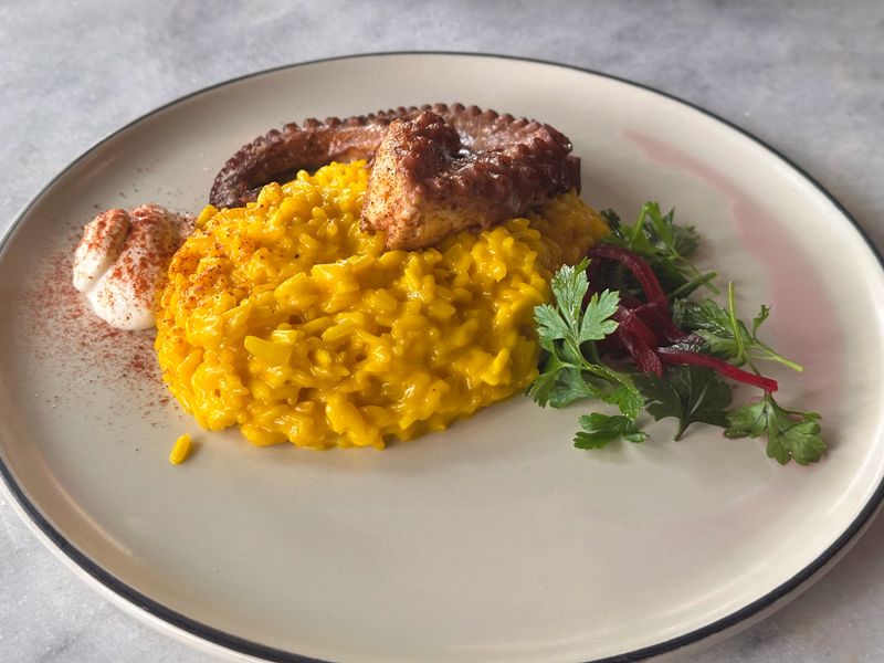 Elegant close-up of a fine dining seafood dish featuring grilled octopus with creamy saffron risotto, paprika yogurt, and fresh herbs. The vibrant yellow rice contrasts beautifully with the grilled octopus, highlighting the sophistication and artistry of modern Mediterranean cuisine.