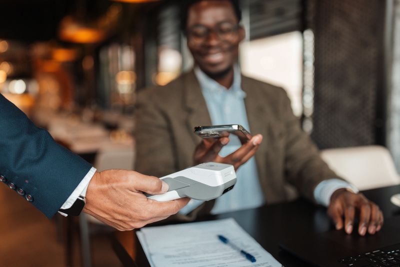 A hand offers a payment device to the man as he holds his phone, depicting the seamless and user-friendly payment processes in contemporary cafes.