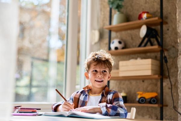 A young boy happily drawing at a table near a window.