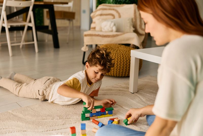 Young boy and his mother bonding while creating structures with wooden toy blocks on a living room rug, fostering development and happy family moments at home