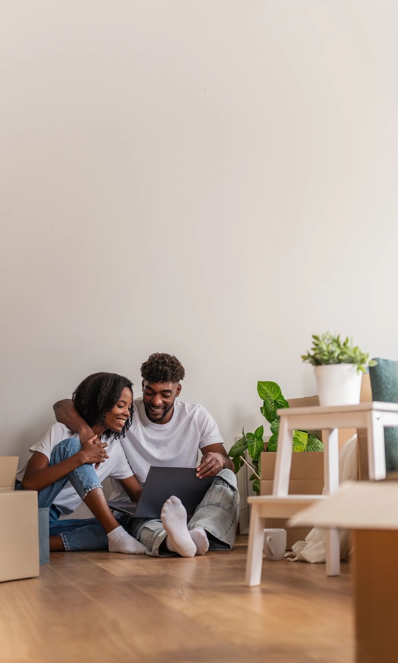 Couple sitting on the floor surrounded by moving boxes, using a laptop in their new home. Cozy atmosphere with plants, couch, and natural light symbolizing a fresh start and togetherness