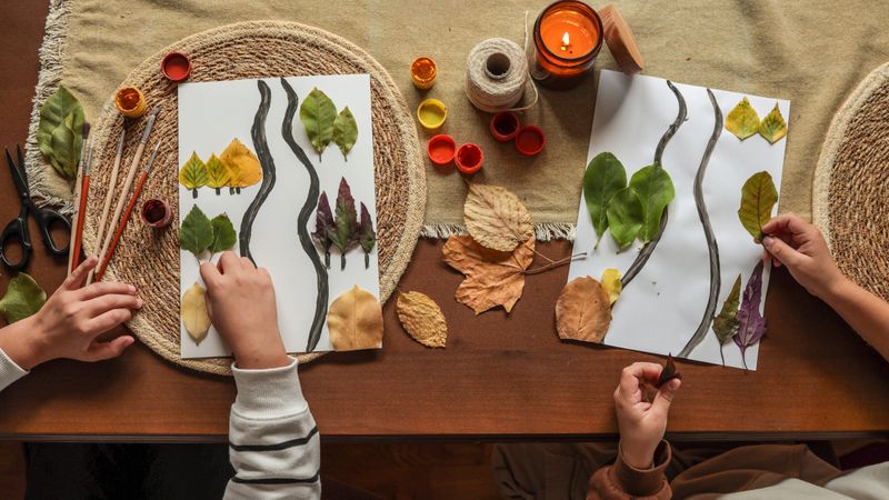 Children are engaged in a creative art project, using leaves and paint to create artwork on paper, with a cozy, autumnal atmosphere.