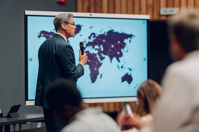 Mature adult businessman speaking at a conference, presenting a global network strategy on a large screen to an audience