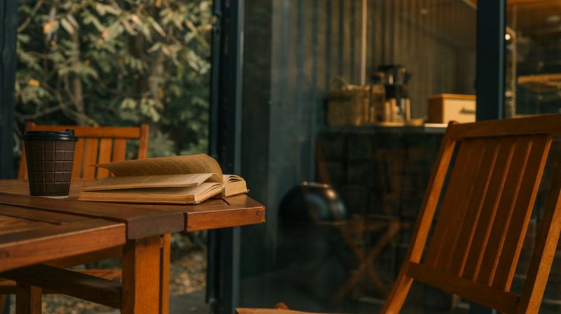 A warm, inviting scene of a book and coffee cup on a wooden table, with a blurred background of a room and nature outside.