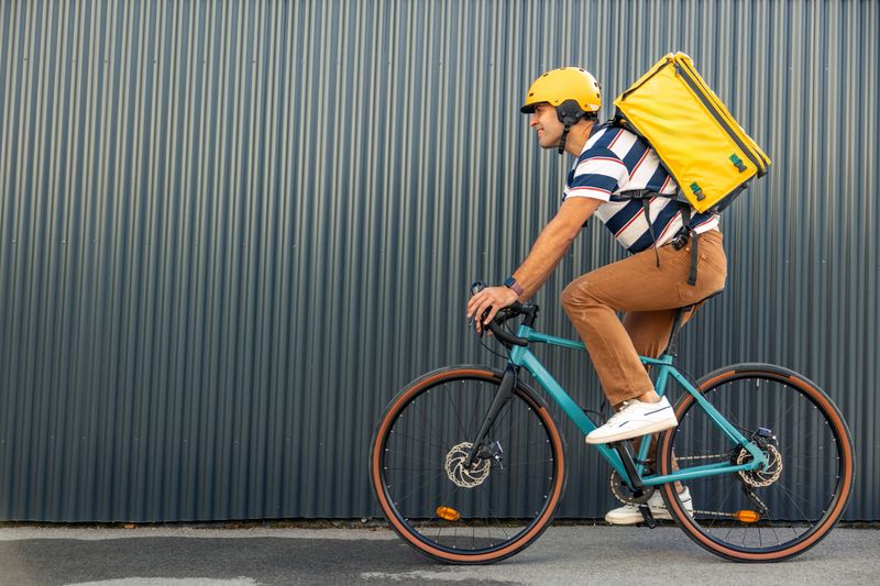 Hispanic male courier wearing a yellow helmet and a delivery bag on his back, biking along an urban wall, performing a delivery service for the gig economy