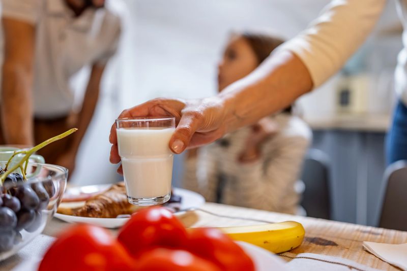 Family breakfast scene with a hand pouring milk into a glass at a sunlit table, featuring tomatoes, bananas, grapes, and pastries.