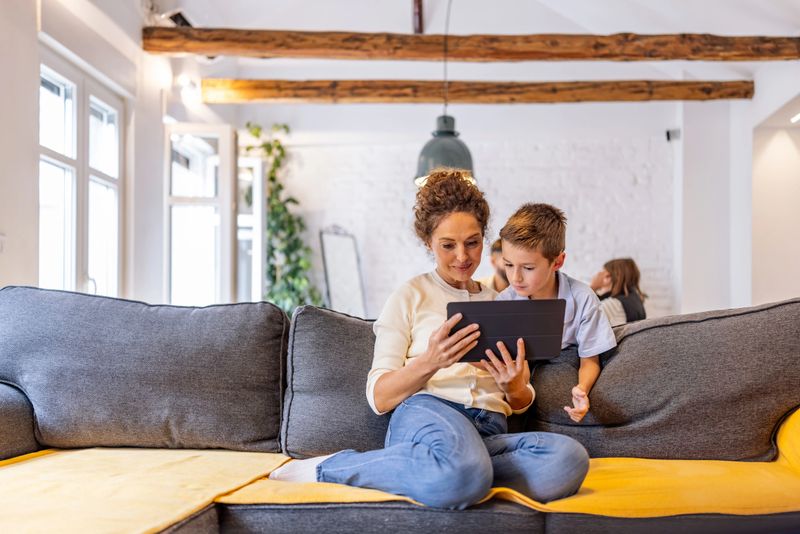 A mother and her young son sit close together on a gray and yellow sectional, sharing a tablet in a bright, stylish living room with exposed wooden beams and greenery.