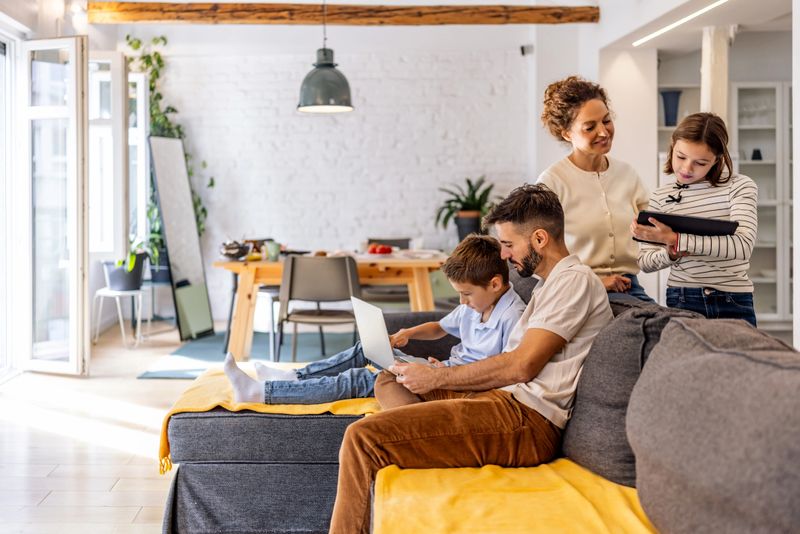A warm family scene in a modern home: father helps son on a laptop while mother and daughter arrive with supplies, capturing togetherness, learning, and everyday life in a comfortable, sunlit living space.