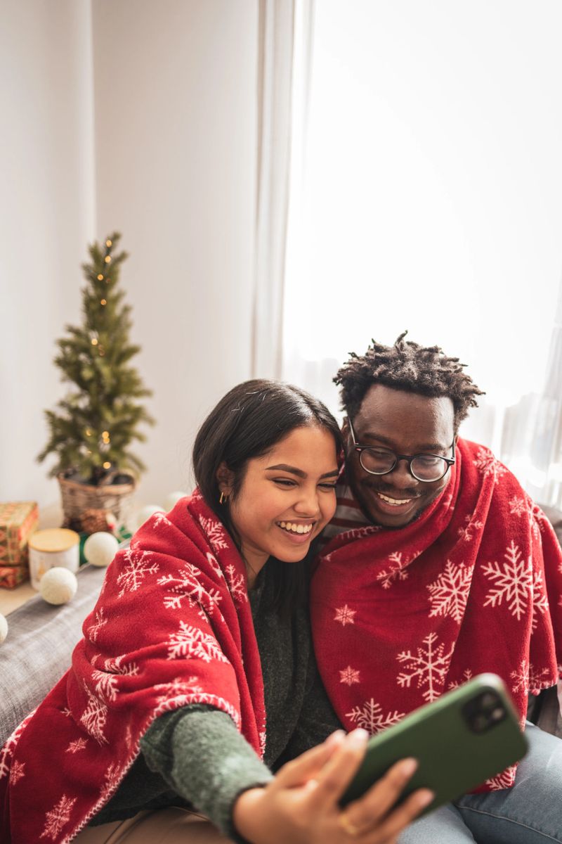 Happy diverse couple wraps themselves in a festive blanket, taking a joyful selfie on their smartphone during christmas