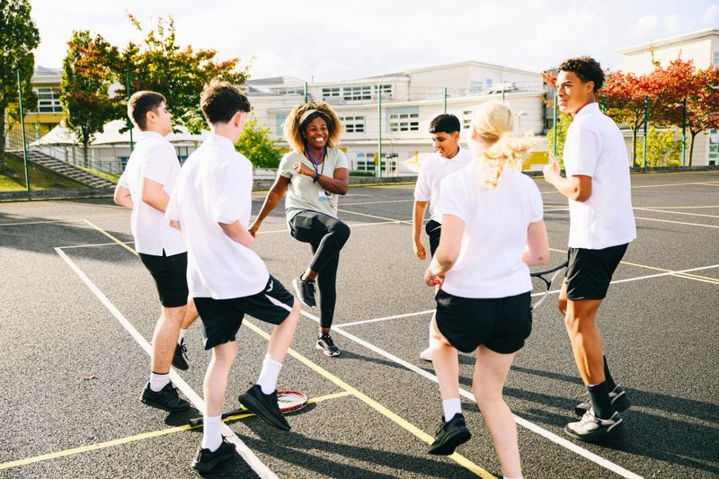 Mature female physical education teacher with a group of five students doing high knees on tennis court before PE lesson
